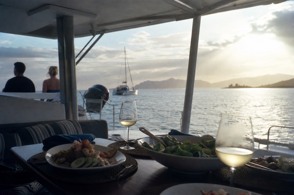 Table set with plated food and wine glasses on the shaded aft deck of a catamaran with the sea blurred beyond the rail