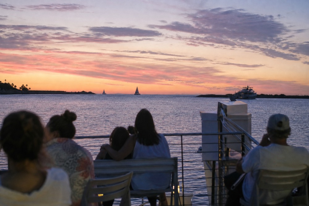 Wide deck view of seated passengers facing west as the light turns warm and low across a calm channel
