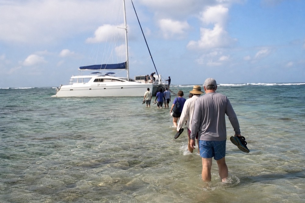 A line of people wading calf-deep in clear water toward the bow step of a beached catamaran, shoes held above the surface