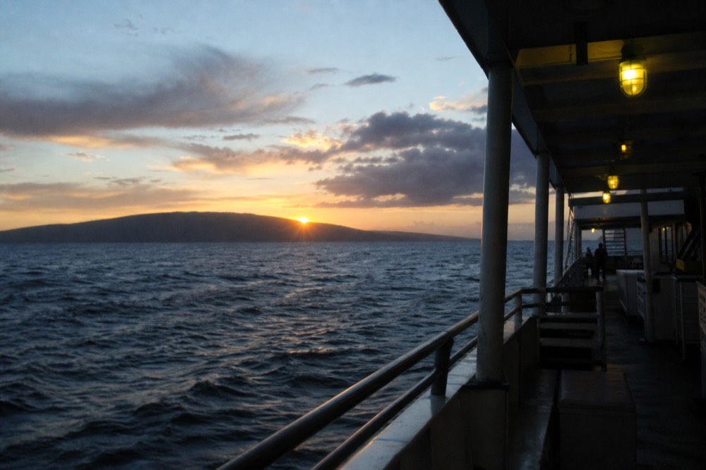 A large low island silhouette filling the western horizon with the sun just above its ridgeline and a dark channel in between