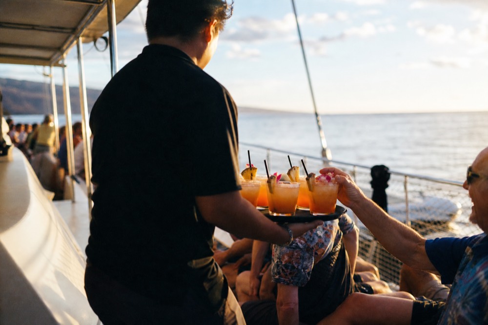A crew member carrying a tray of tropical cocktails along a narrow boat deck, guests reaching for glasses in warm side light