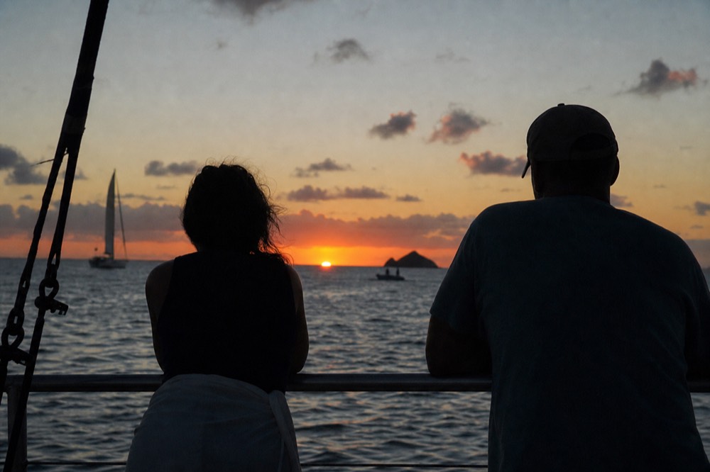 The last sliver of sun touching the sea horizon seen between two figures leaning on a boat rail, everything else in silhouette