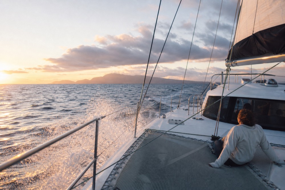 Spray bursting over the windward hull of a sailing catamaran with a guest in a light jacket laughing on the netting