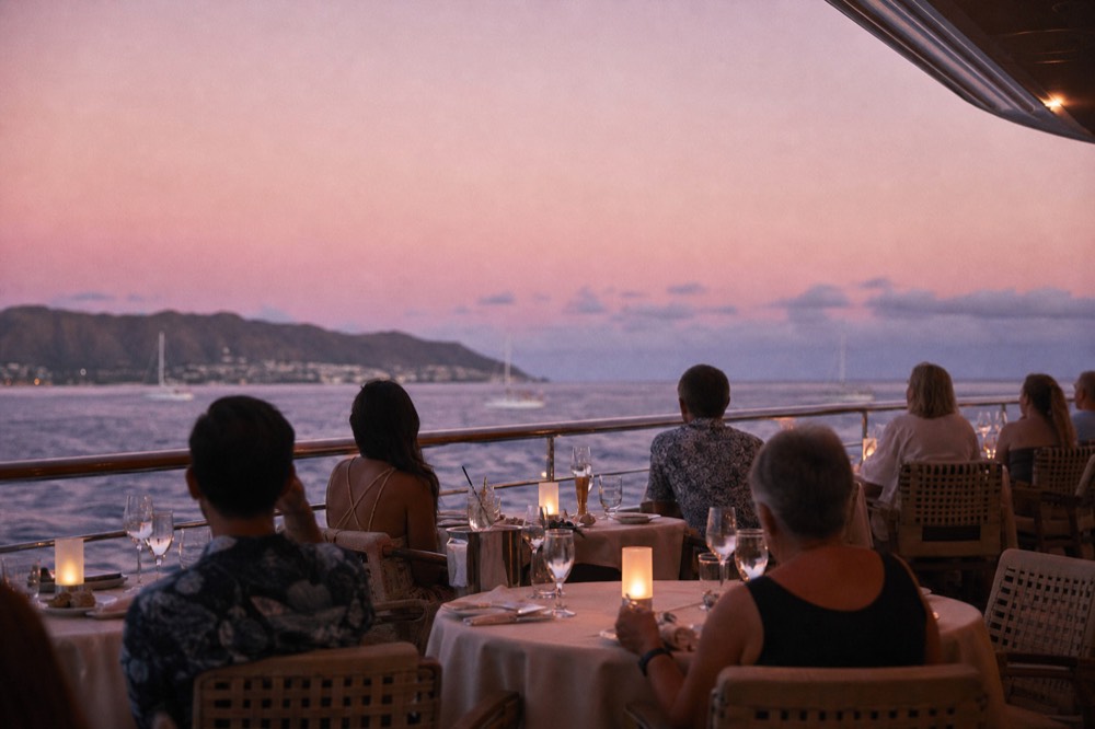 Guests in relaxed resort clothes seated at set dinner tables on an open upper deck before sunset