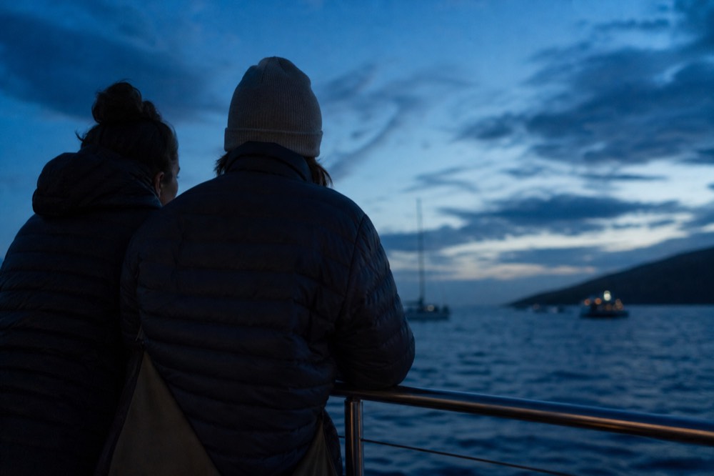 Two people in warm layers standing close together at a boat rail in blue-hour light after the sun has gone