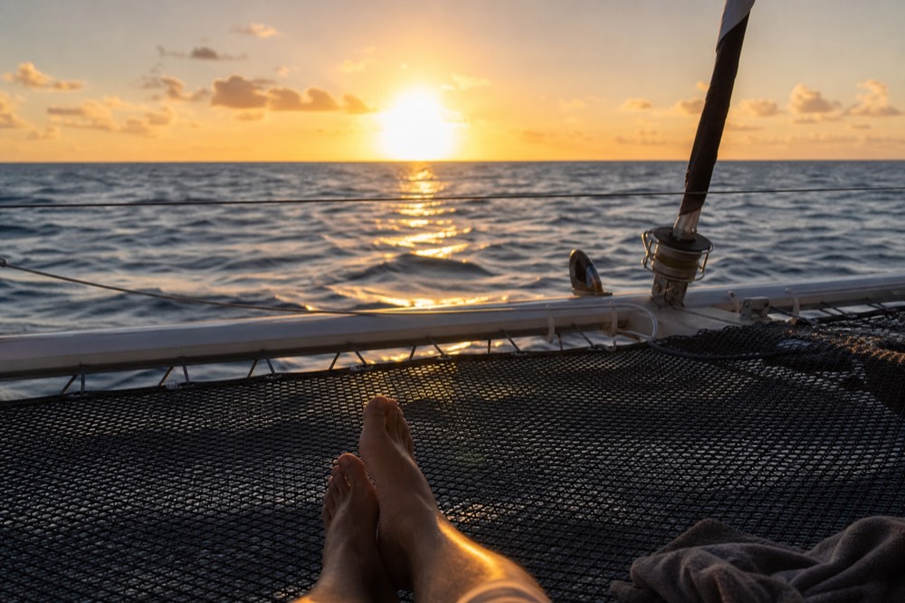 Bare feet on a boat trampoline net with open ocean and low orange light directly ahead and no shoreline anywhere in the frame