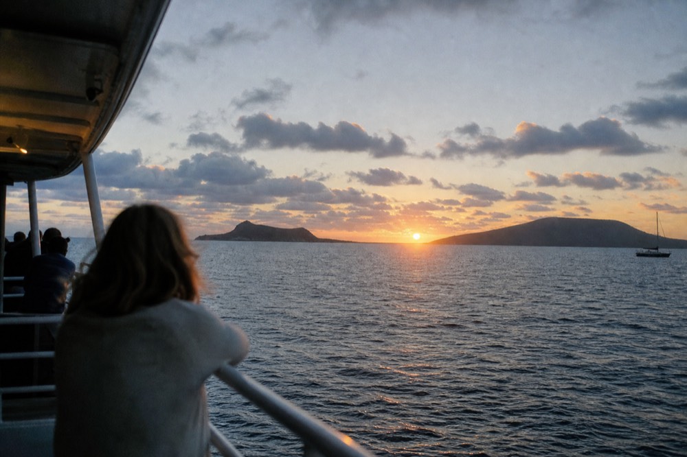 Sun dropping into the notch between two low island silhouettes seen from a boat rail in midsummer light