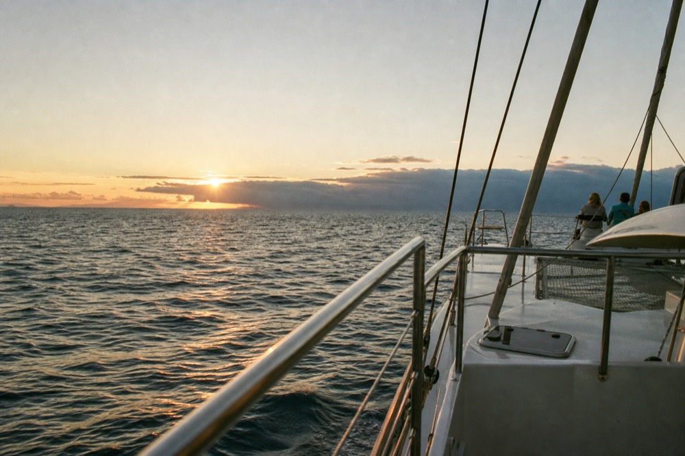 Low bank of trade wind cloud sitting on the horizon at sunset with light spilling over the top and a boat deck rail in the near foreground