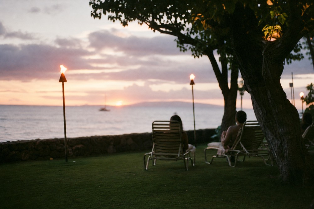 Resort lawn set with chairs facing the water at dusk with torches lit along the edge and the ocean beyond a low stone wall